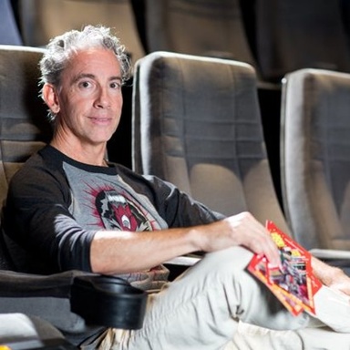 Man sitting in movie theatre holding comic book, smiling at camera.